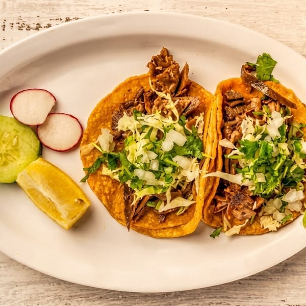 Two street tacos with meat, cabbage, and cilantro, served with cucumber, radish, and lemon.